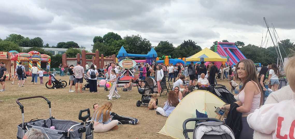 Bouncy castles and rides in the background with a large group of people in the foreground attending the party in the park event at hatfield peverel